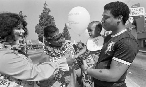 Two women dressed as clowns talking with a man and a baby at a parade, Los Angeles, 1982