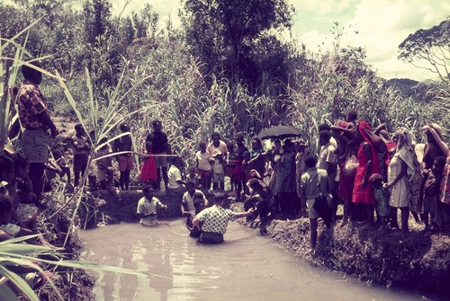 Protestant missionary helps a convert enter a pond for baptism, surrounded by group of onlookers