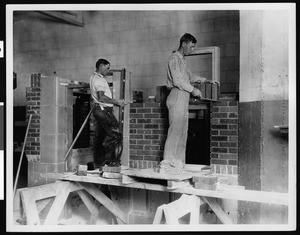 Two students laying bricks and setting door frames on a model home, ca.1920-1929
