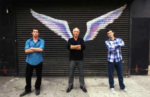 Three unidentified men posing with arms crossed in front of a mural depicting angel wings