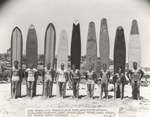 Club surfers standing in front of boards: Alex Hokamp, Rich Thompson, Fred Hunt, Dave "Buster" Steward, Alex "Pinky" Pedemonte, Tommy Roussel, Blake Turner, Harry Murray, Don "Mighty Bosco" Patterson on beach in front of Dream Inn