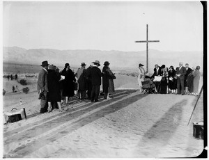 People congregating around a cross and piano for a funeral in the desert
