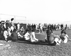 A bunch of people watch a softball game in progress, as a woman approaches the plate