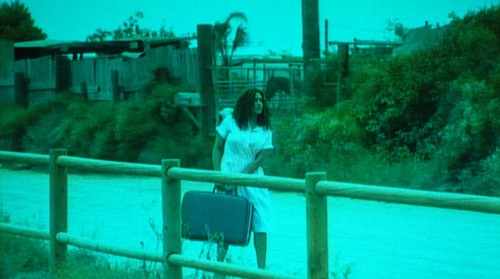 Border stories working title from one side to another: film still of woman carrying suitcase down a road