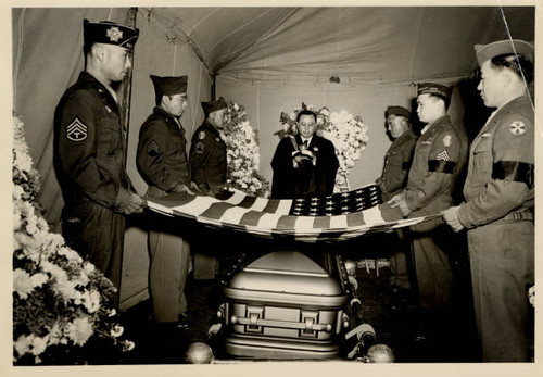 Japanese Americans in military uniforms hold American flag over casket while priest gives service