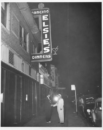 Two unidentified men stand shaking hands on sidewalk underneath Elsie's neon sign