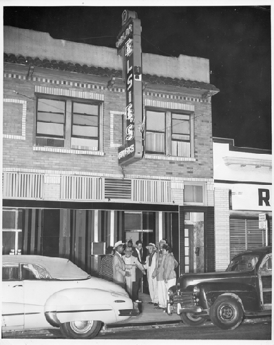 Patrons stand on sidewalk of Elsie's with automobiles parked in front