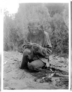Old Havasupai Indian man crouching on the ground, rolling a cigarette, ca.1900