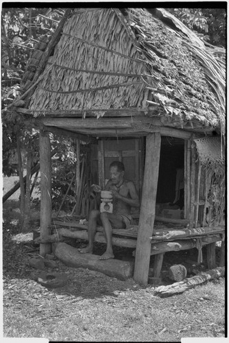 Carving: M'lapokala carving a wooden bowl with a stand, probably for tourist trade, he sits on veranda of a small house