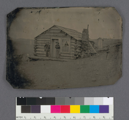 [Two men in front of a log cabin.]