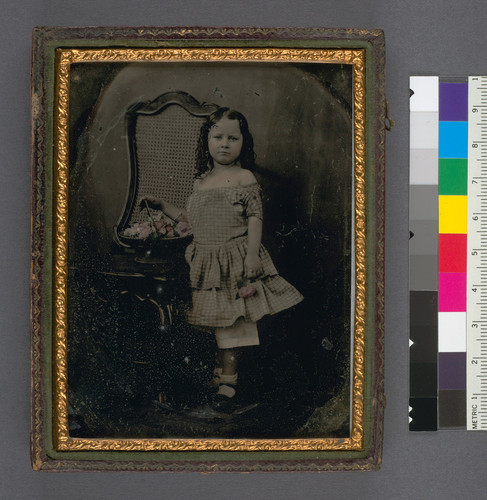 [Little girl with a basket of flowers, standing next to a chair.]