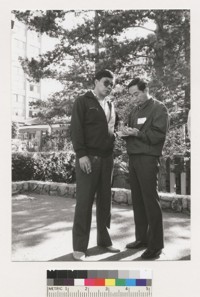 Two unidentified men standing, one is writing something
