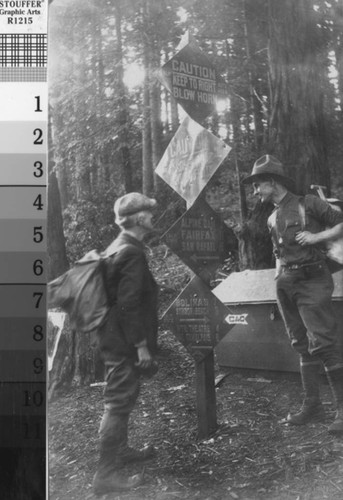 Hiker and Ranger standing on trail wearing backpacks standing next to signpost with warning and directional signs