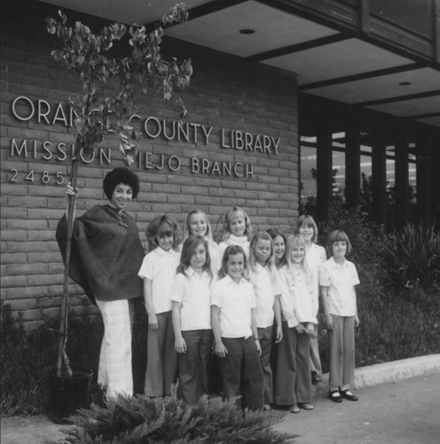 [Girls from Mission Viejo Bluebird group and a woman standing with potted tree in front of Orange County Library Mission Viejo Branch, 1976 photograph]