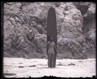 Surfer and Olympic swimmer Duke Kahanamoku standing on a beach with a surfboard, Los Angeles, 1920s