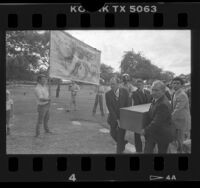 Two men with anti-abortion banner watch as bearers carry casket of aborted fetuses to gravesite in Los Angeles, Calif., 1985