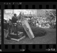 UCLA student body President Robert Michaels standing beside coffin marked "University of California," as he speaks to crowd at budget cut protest, 1967