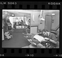 Dentist Dr. Scott Jacks examining one patient while two other patients play video games in waiting area, South Gate, Calif., 1982