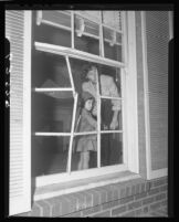 Mrs. William Gude and daughter Wendy look through shattered window, Brentwood (Los Angeles), 1950