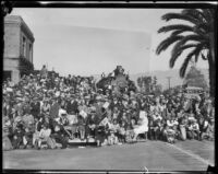 Rose Parade spectators next to building and train engine, Pasadena, 1930