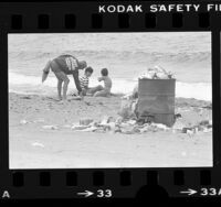 Lifeguard picking up litter on trash strewed beach in Los Angeles, Calif., 1979