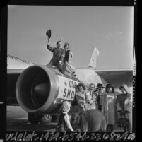 Bob Hope perched on jet engine as other entertainers gather before leaving on Christmas USO tour, 1964