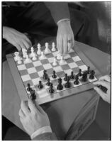 Top view of a chess board and both of the players' hands, one of the players is moving the white knight, Los Angeles