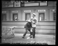 Chico and Groucho Marx standing at ticket windows during filming of scene for "A Day at the Races", Los Angeles, 1937