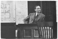 Murder suspect Robert S. James sitting on the witness stand with a map of his house behind him, Los Angeles, 1936