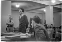 Man testifying (?) during unidentified hearing in courtroom with model cars on table