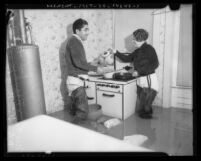 Couple wearing waders, cooking at stove in flooded kitchen, 1937 flood Long Beach, Calif