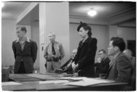 Man and woman testifying (?) during unidentified hearing in courtroom with model cars on table