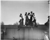 Men perched on a fence at Spaulding Field, UCLA, to photograph the Bruins football team on opening day of the season, Los Angeles, 1937