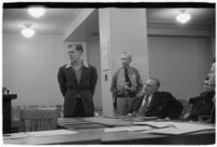 Man testifying (?) during unidentified hearing in courtroom with model cars on table