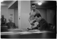 Man testifying (?) during unidentified hearing in courtroom with model cars on table