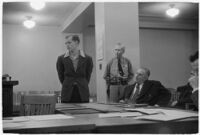 Man testifying (?) during unidentified hearing in courtroom with model cars on table