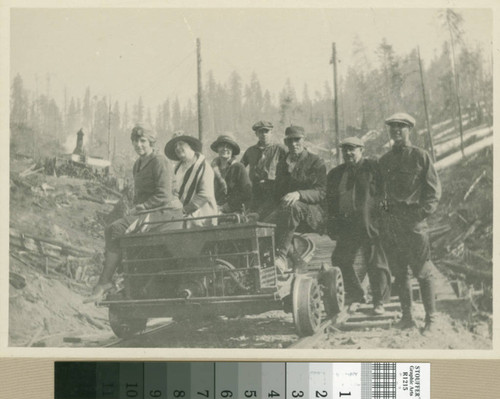 [4 men, 3 women posed on rail car in logged forest]