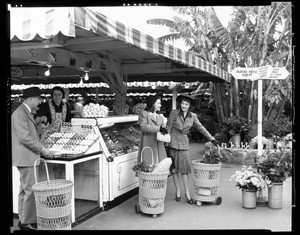 A man gets his change from the woman working at the egg counter at the outdoor Farmers Market, while two young women with wicker shopping carts stand nearby