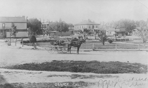 Man and young boy in four-wheeled, horse-drawn carriage, posed by the Plaza in Orange, California, 1890