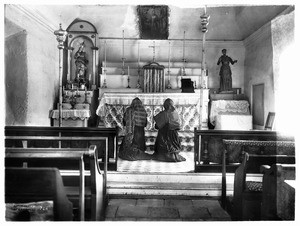 Interior view of Mission San Juan Capistrano, showing two worshippers kneeling in front of the altar, ca.1900