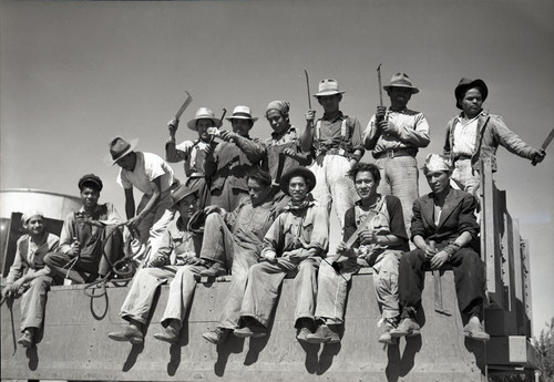 Fourteen Mexican workers sitting on back of flatbed truck holding hoes