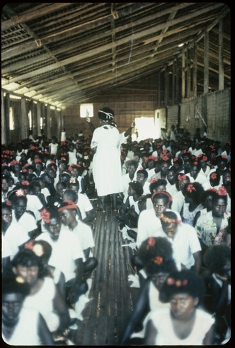 Group seated in a building, with Holy Mama standing in center; most are in white dress with red flowers in hair