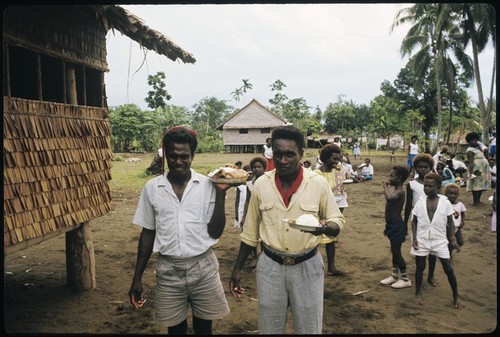 Two men carrying plates with food. Women and children in back