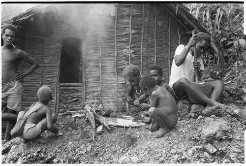 Taaboo lighting pipe in front of men's house with kids; another man in white shirt is pounding taro for pudding