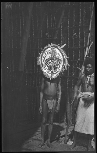 Kaimari village men standing by a fire pit inside a house; one is wearing a mask