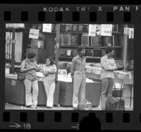 Four people waiting at bus stop as Richard Nixon's resignation address plays on television in electronics store behind them, Los Angeles, Calif., 1974