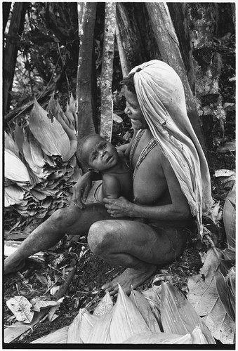 Pig festival, pig sacrifice, Tsembaga: woman with flattened leaves for sacrifice house thatching, holds infant