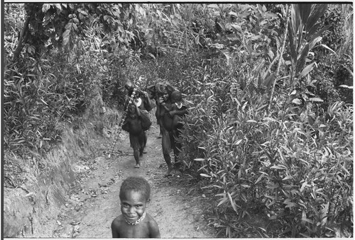 Women carry netbags and children on trail, also bamboo water container