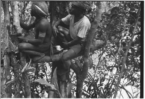 Bridge-building: men sit on frame lashed with vines, one man holds steel axe