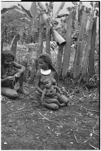 Women sit together, one working with string, the other holding infant